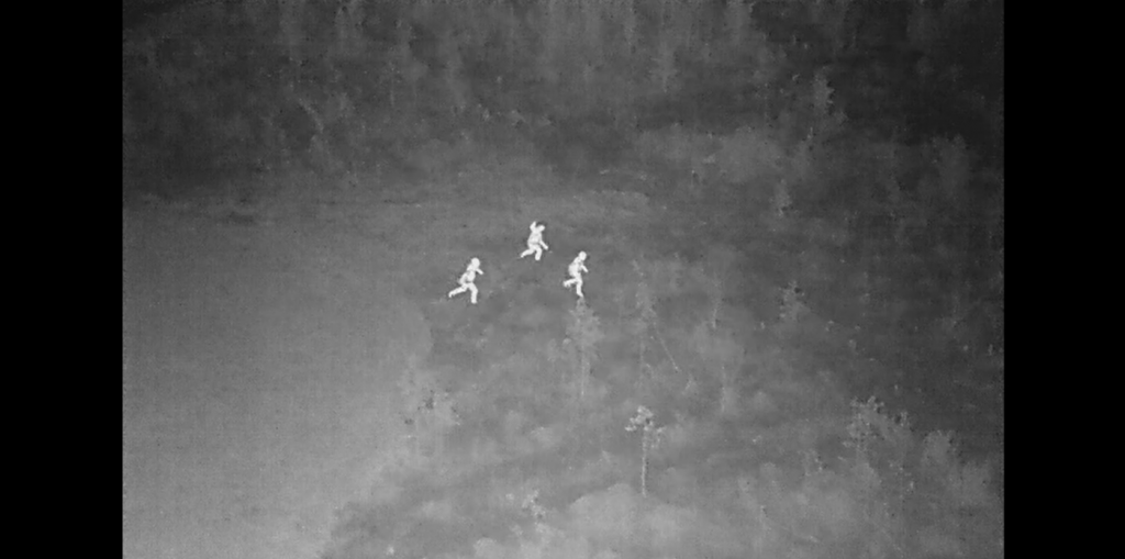 Thermal camera photo, which shows people running in the forrest at night.