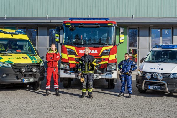 Valokuvassa ensihoitaja, palomies ja poliisi sekä ambulanssi, paloauto ja poliisiauto.